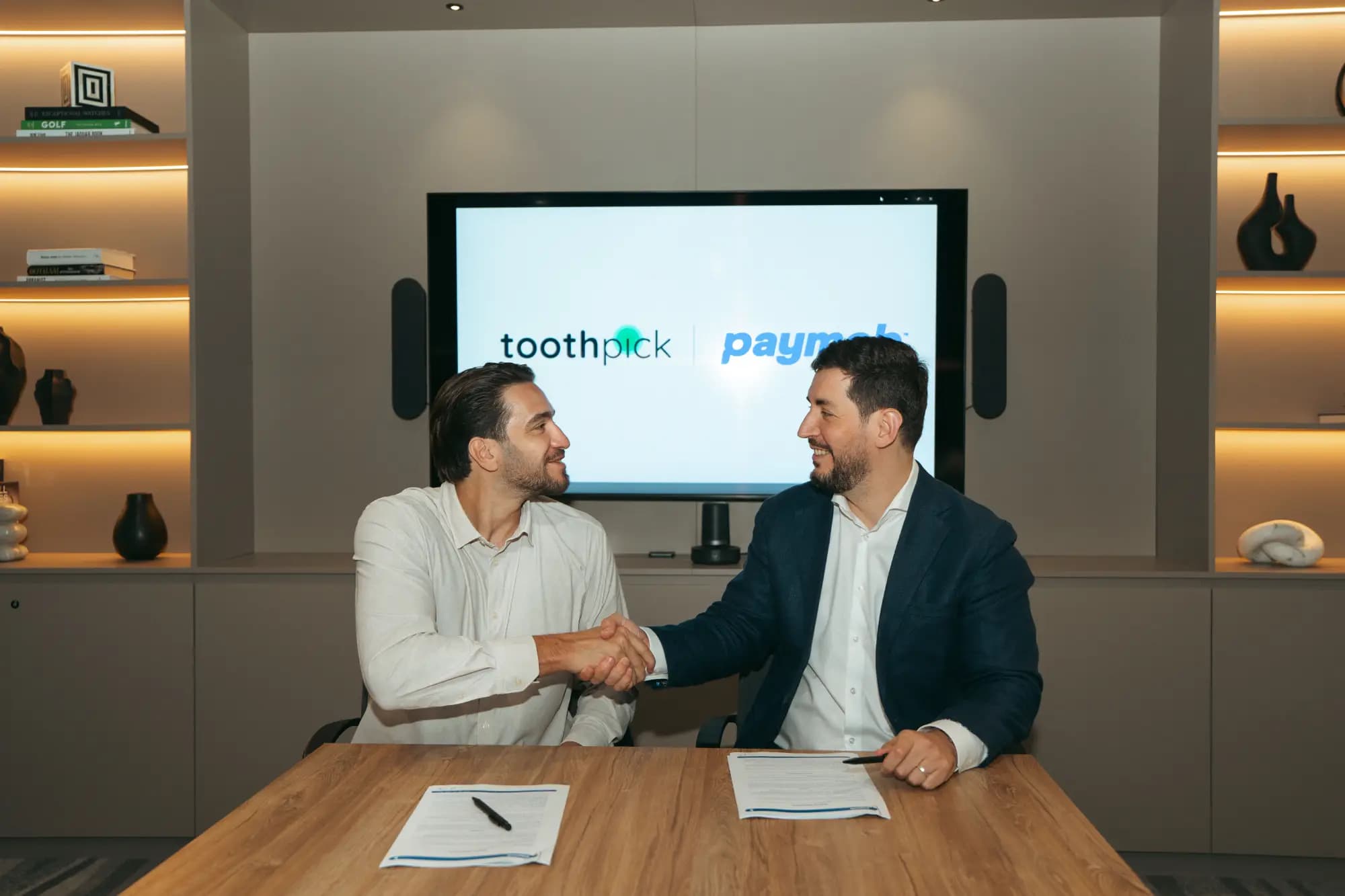 Two representatives shaking hands across a table in front of the Toothpick and Paymob logos