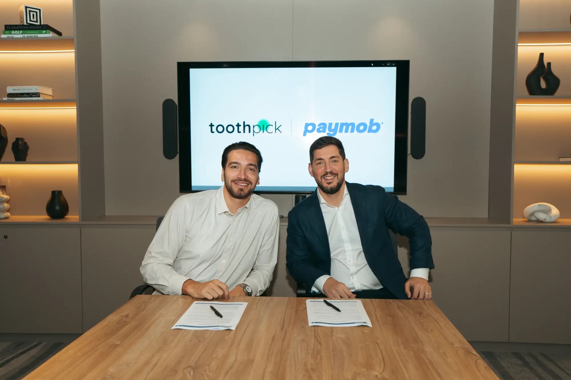 Two representatives seated at a table with documents and pens, in front of a screen showing the Toothpick and Paymob logos