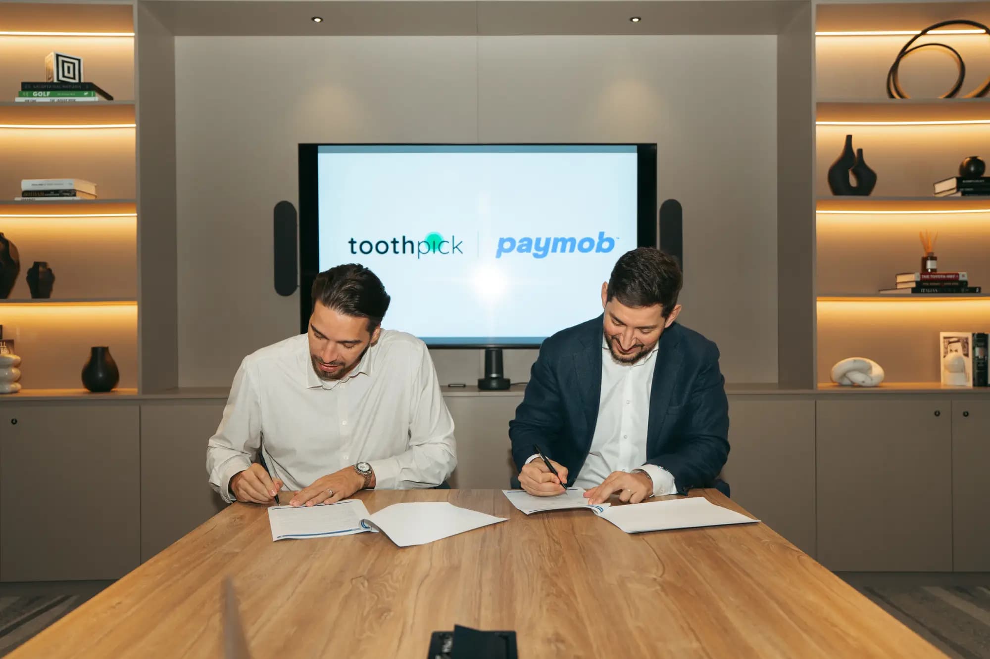 Two representatives signing documents at a table, with the Toothpick and Paymob logos on the screen behind them